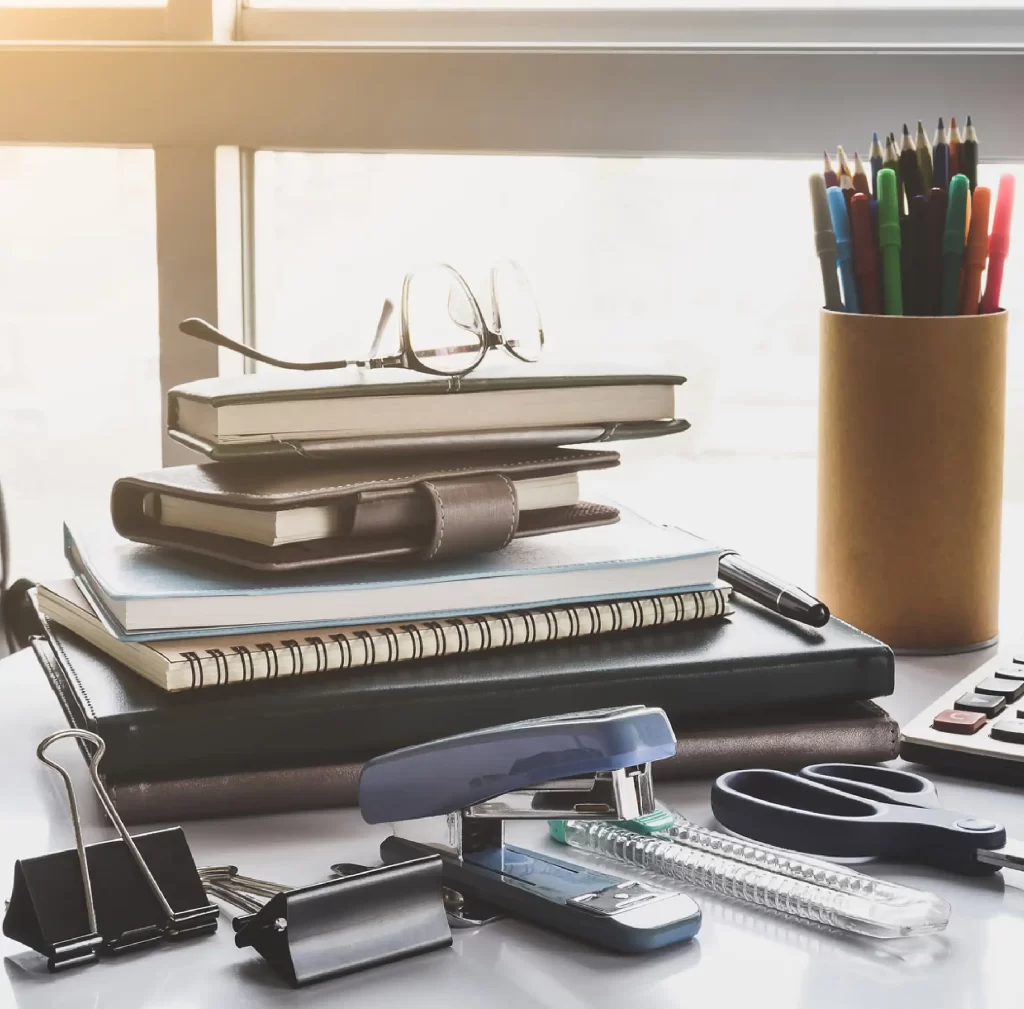 A variety of stationery items are scattered across a desk, including notebooks, pens, pencils, scissors, a stapler, and paper clips.