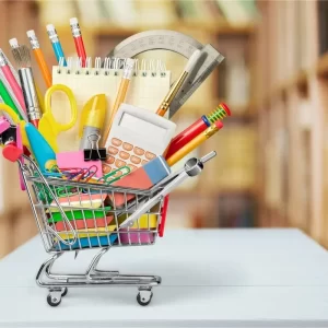 A miniature shopping cart filled with office stationery items, including pens, pencils, scissors, notepads, a calculator, and other supplies, set against a blurred background.